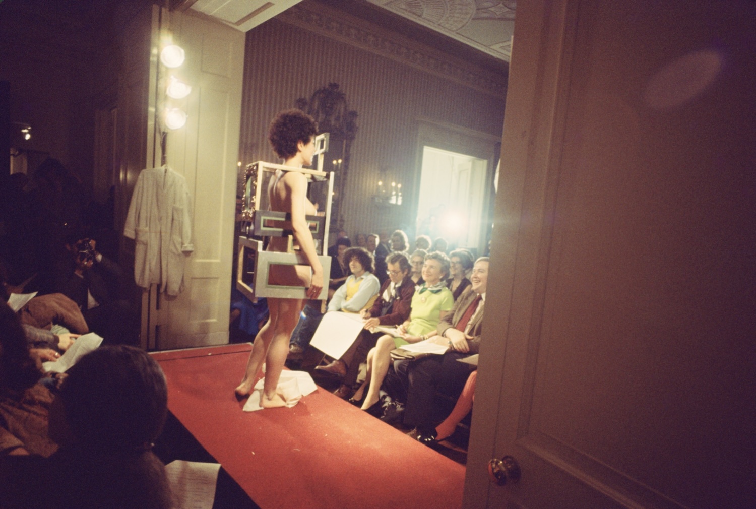 A naked woman with short curly hair stands within what looks like a boxy dress made of matting frames on a runway in front of an audience.