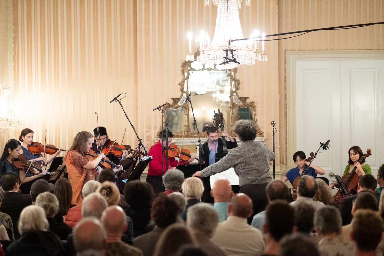 A string ensemble plays before a conductor and a crowd in a baroque ballroom.