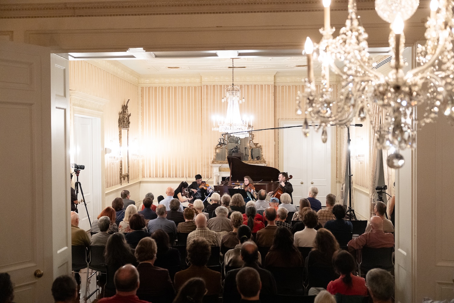 A string ensemble plays before a crowd in a baroque ballroom.