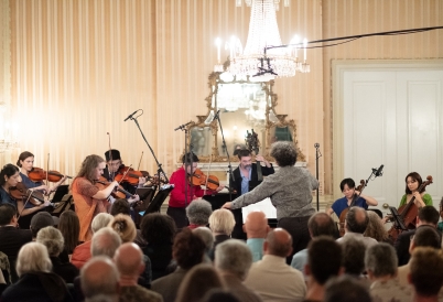 A string ensemble plays before a conductor and a crowd in a baroque ballroom.