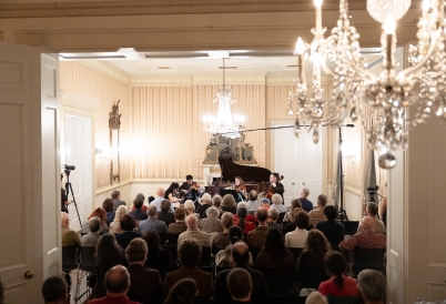 A string ensemble plays before a crowd in a baroque ballroom.