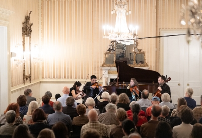 A string quartet plays in front of a small audience in a baroque ballroom.