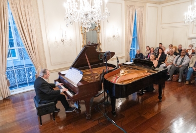 A man and a woman play at two pianos opposite each other in front of an audience in a baroque ballroom.