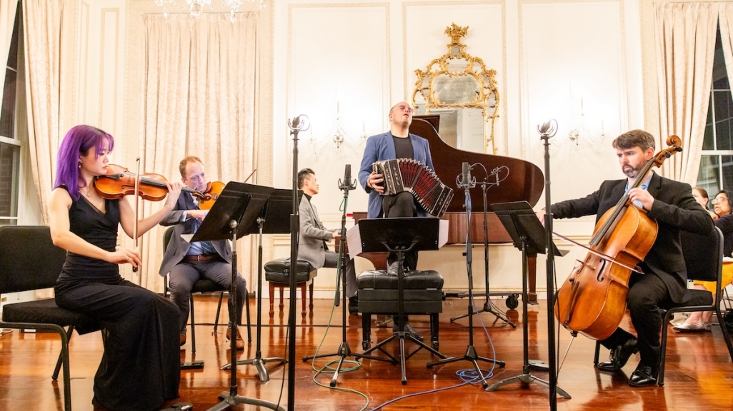 A chamber music ensemble, with accompanying accordion player, perform in a ballroom.